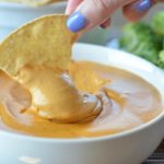 A vertical shot of pretty lady fingers with blue nails dipping a tortilla chip into a bowl of cashew queso.