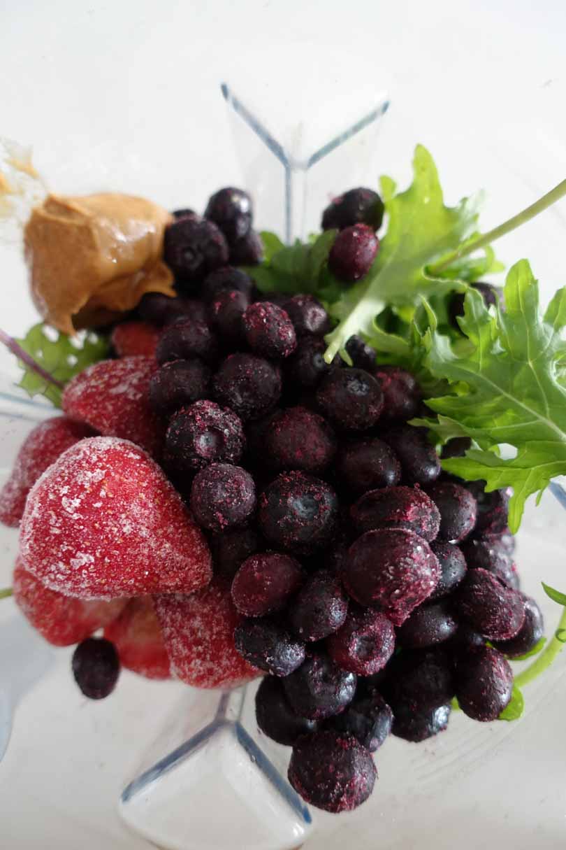 Frozen strawberries blueberries peanut butter and greens in a Vitamix container.